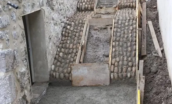 Vue d'ensemble du chantier en cours d'exécution : gestion de la pente, alignement des marches en pierre et préparation des coffrages en pied de structure pour couler les dernières sections de galets scellés. »